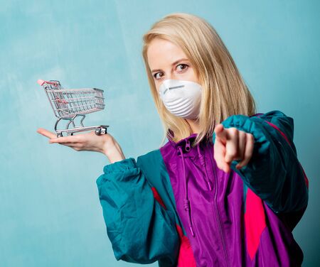 Style blonde woman in 90s clothes and face mask with shopping cart on blue background