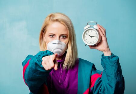 Blonde woman in face mask and 90s style clothes with alarm clock on blue background