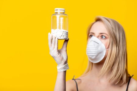 Blonde woman in face mask and gloves holds jar with pills on yellow backgroundの写真素材