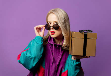Style blonde in 80s windbreaker and roud sunglasses with gift box on purple background