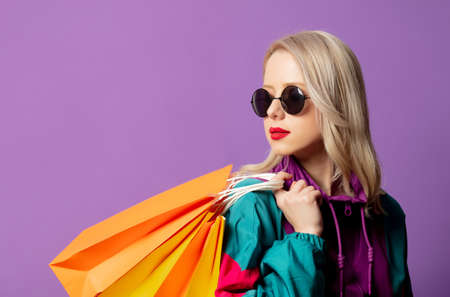 Style woman in 80s windbreaker and roud sunglasses hold shopping bags on purple background