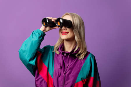 Style woman in 80s windbreaker and roud sunglasses hold binoculars on purple background