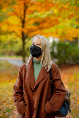 Style blonde in face mask and coat on autumn park with maples on background