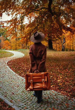 Style girl in brown coat with suitcase in autumn parkの写真素材
