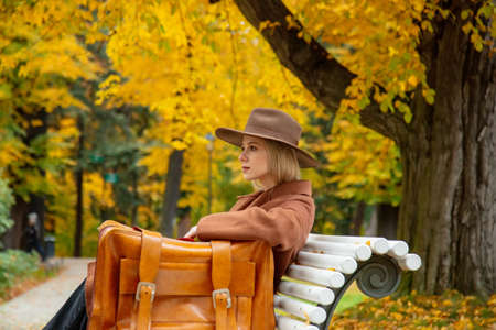 Style girl in brown coat with suitcase sitting on a bench in autumn parkの写真素材