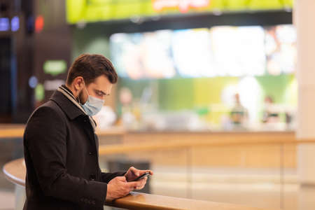 Guy in face mask and coat is using mobile phone in city mall in Pandemic time