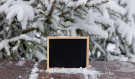 Blackboard on wooden table with snow next to pine treeの写真素材