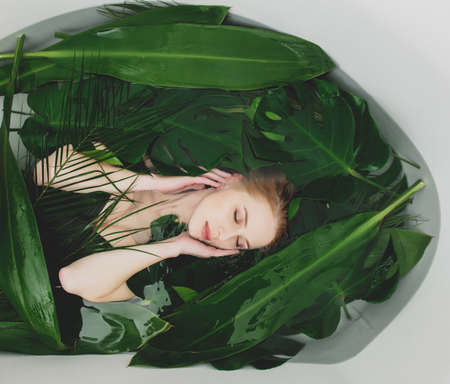 Beautiful white woman lying in bathtub with palm leaves around