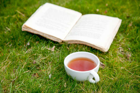 Old book with cup of tea on green grass in springの写真素材