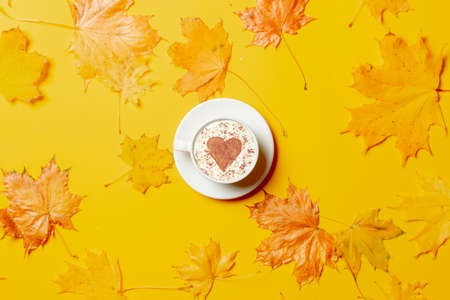 Cup of coffee with heart shape and maple leaves around on yellow background.の写真素材