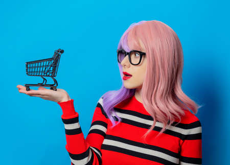 Girl with purple hair and red sweater hold black shopping cart on blue backgroundの写真素材