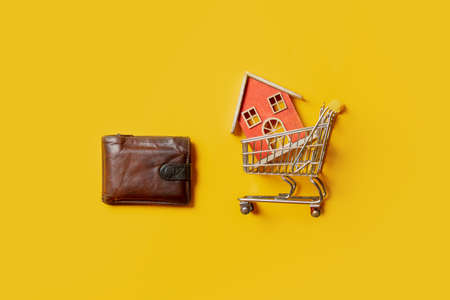 Toy house in supermarket cart and wallet on yellow background. Top viewの写真素材