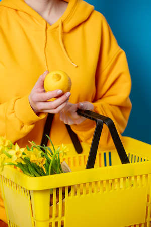 Stylish female in yellow hoodie with lemon, basket and flowers on blue backgroundの写真素材