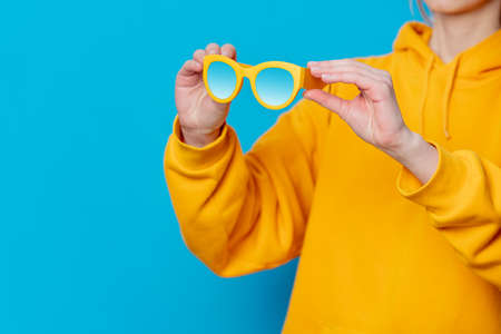 Female in yellow hoodie holding sunglasses in blue backgroundの写真素材
