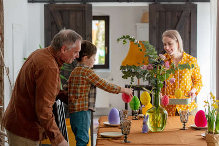 Grandpa, grandson and mother are getting ready for Easter dinner. At the table in the house, beside a decoration of paper eggs.の写真素材