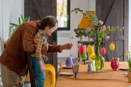 Grandpa and his grandson decorate the bouquet of flowers with paper Easter eggs. At home during the holidayの写真素材