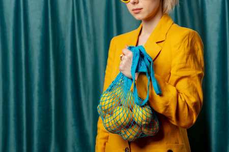Stylish ukrainian woman in yellow sunglasses and jacket with lemons in net bag on green curtains backgroundの写真素材