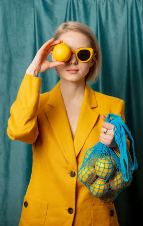Stylish ukrainian woman in yellow sunglasses and jacket with lemons in net bag on green curtains backgroundの写真素材