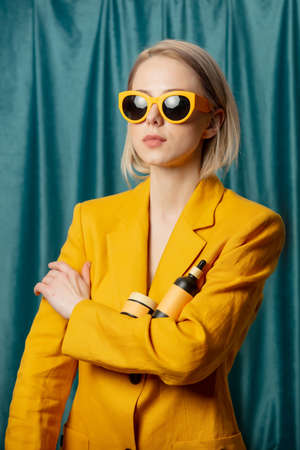Stylish ukrainian woman in yellow sunglasses and jacket with cosmetic bottle on green curtains backgroundの写真素材