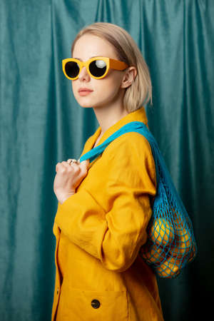Stylish ukrainian woman in yellow sunglasses and jacket with lemons in net bag on green curtains backgroundの写真素材