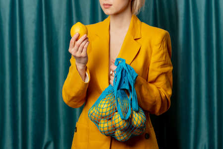Stylish ukrainian woman in yellow sunglasses and jacket with lemons in net bag on green curtains backgroundの写真素材