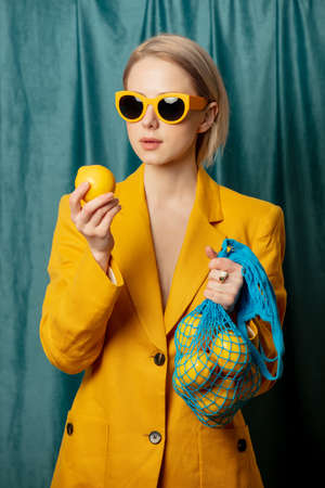 Stylish ukrainian woman in yellow sunglasses and jacket with lemons in net bag on green curtains backgroundの写真素材