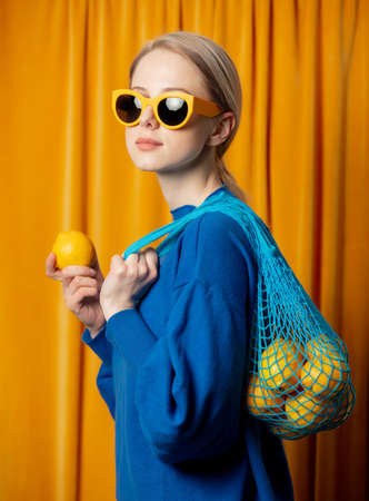 Stylish ukrainian woman in yellow sunglasses and blue sweater with lemons in net bag on curtains backgroundの写真素材