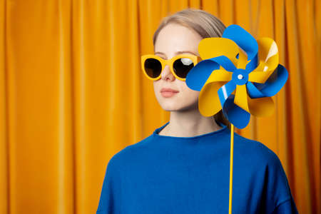 Stylish ukrainian woman in yellow sunglasses and blue sweater with pinwheel in ukrainian flag colors on curtains backgroundの写真素材
