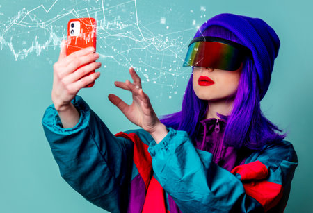 Stylish girl in cyber punk glasses and 80s tracksuit using mobile phone on blue backgroundの写真素材