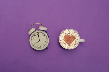 Cup of cappuccino with cinnamon heart design, vintage white alarm clock on a purple background, top viewの写真素材