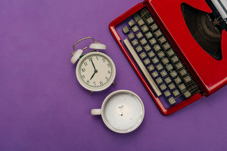 Vintage white alarm clock next to a red retro typewriter and a cup of coffee on a purple background, top viewの写真素材