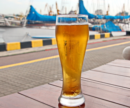 A glass of cold beer standing on a red table against the backdrop of a yachtの写真素材