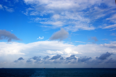 White clouds of various shapes against the blue sky and the sun above the surface of the ocean.の写真素材