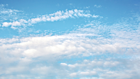 White clouds of various shapes against the blue sky and the sun above the surface of the ocean.の写真素材