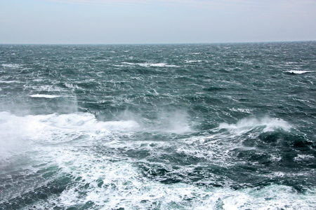 Storm waves in the world ocean. Kind of waves, crests, splashes, foam against the background of the sea horizon and the blue sky and clouds. A view of storm waves from the ship's side.の写真素材