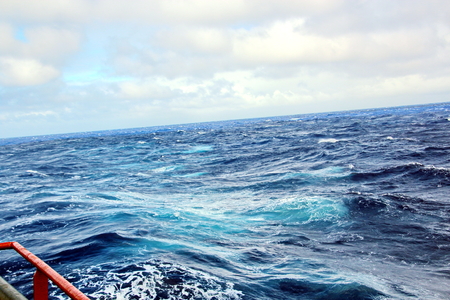 Storm waves in the world ocean. Kind of waves, crests, splashes, foam against the background of the sea horizon and the blue sky and clouds. A view of storm waves from the ship's side.の写真素材