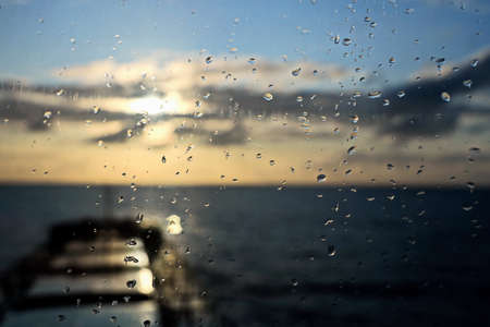 View of the ocean and sky through raindrops on the glass of a ship's porthole.