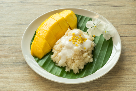Ripe yellow mango and sticky rice with coconut milk cream topping yellow bean served on banana leaf on white plate on wood table.の写真素材