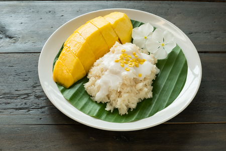 Ripe yellow mango and sticky rice with coconut milk cream topping yellow bean served on banana leaf on white plate on wood table.の写真素材