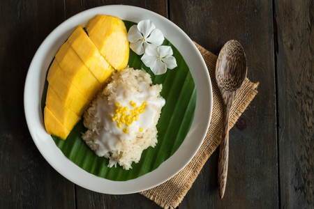 Ripe yellow mango and sticky rice with coconut milk cream topping yellow bean served on banana leaf on white plate on wood table with wood spoon.の写真素材