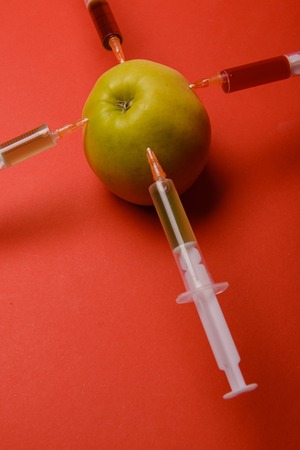 Chemical additives in food or genetically modified fruit concept. Green apple with syringes of chemicals. Isolated on red background.の写真素材