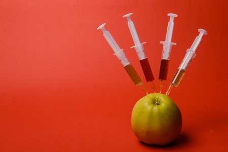 Chemical additives in food or genetically modified fruit concept. Green apple with syringes of chemicals. Isolated on red background.の写真素材