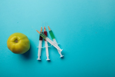 Chemical additives in food or genetically modified fruit concept. Green apple with syringes of chemicals. Isolated on blue background.の写真素材