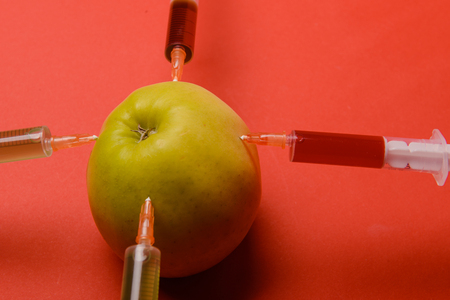 Chemical additives in food or genetically modified fruit concept. Green apple with syringes of chemicals. Isolated on red background.の写真素材