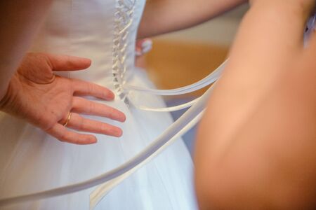 Bridesmaid helping slender bride lacing her wedding white dress, buttoning on delicate lace pattern...