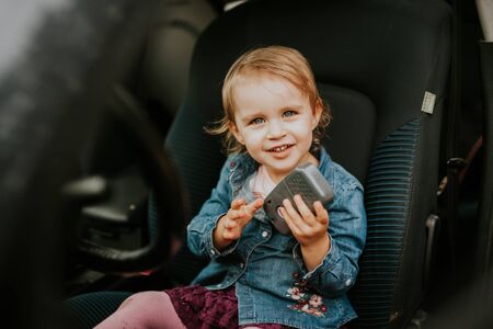 Little girl in a car seat. Holding phone in her hand. Taking driver seat. Car damage. Authentic image.の写真素材