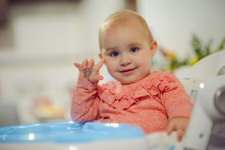 cute blond child sits in a children's chair at the kitchen and eat a juicy peach. Little girl with a piercing look.の写真素材