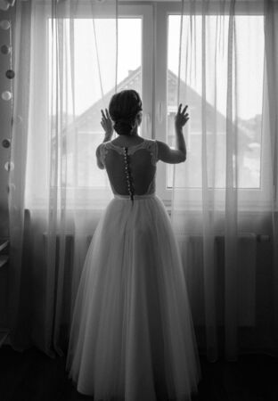 Bridesmaid helping slender bride lacing her wedding white dress, buttoning on delicate lace pattern with fluffy skirt on waist. Morning bridal preparation details newlyweds. Wedding day moments, wear.の写真素材