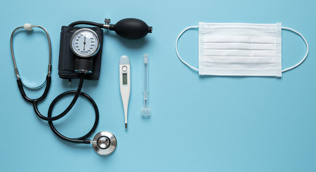 A collection of essential medical tools, including a stethoscope, blood pressure monitor, digital thermometer, and a protective face mask, arranged on a blue surface for healthcare purposes.の素材