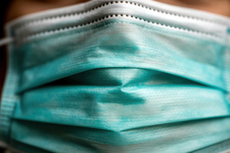 Extreme close-up of a teal blue disposable surgical face mask. The image focuses on the pleated fabric texture with visible stitching along the top edge. The material shows subtle variations in color and light, highlighting its woven structure and folds.の素材